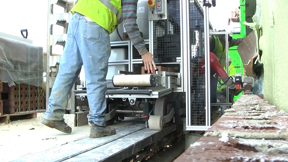 SAM100 uses a laser beam string line to accurately place a brick, while in the foreground a mason tender from Manning Construction adds bricks to the system’s conveyor.
