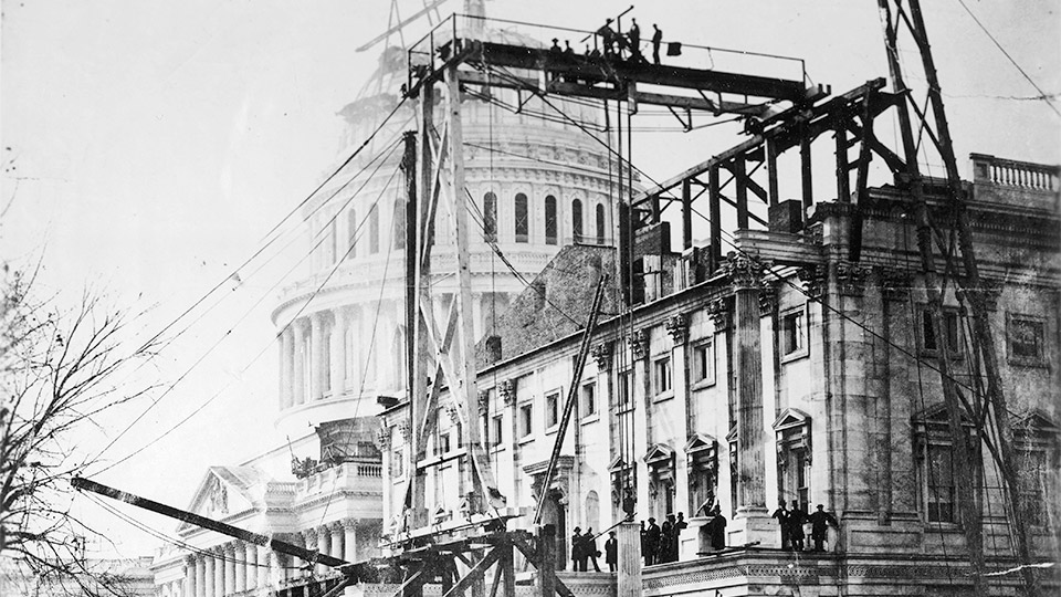 Shown is the east elevation of the Senate Extension, a column shaft is being hoisted into place. The scaffold, gantry and derricks designed to hoist and move the large pieces of stone are engineering marvels. Note the last of safety requirements — lots of people watching under the rigging and no hard hats or no overhead protection! c.1864.